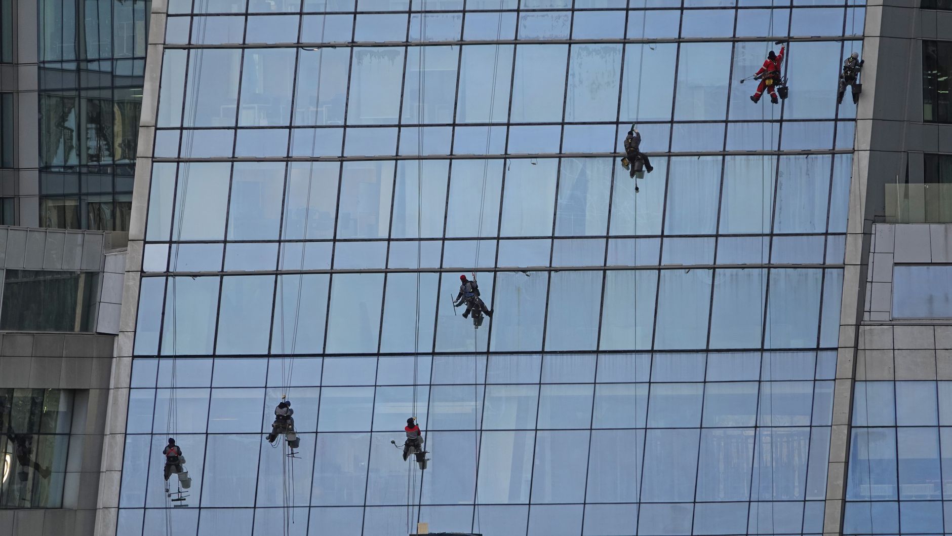 Rope-access workers in harnesses suspended on a glass office building facade, reflecting sky and windows.