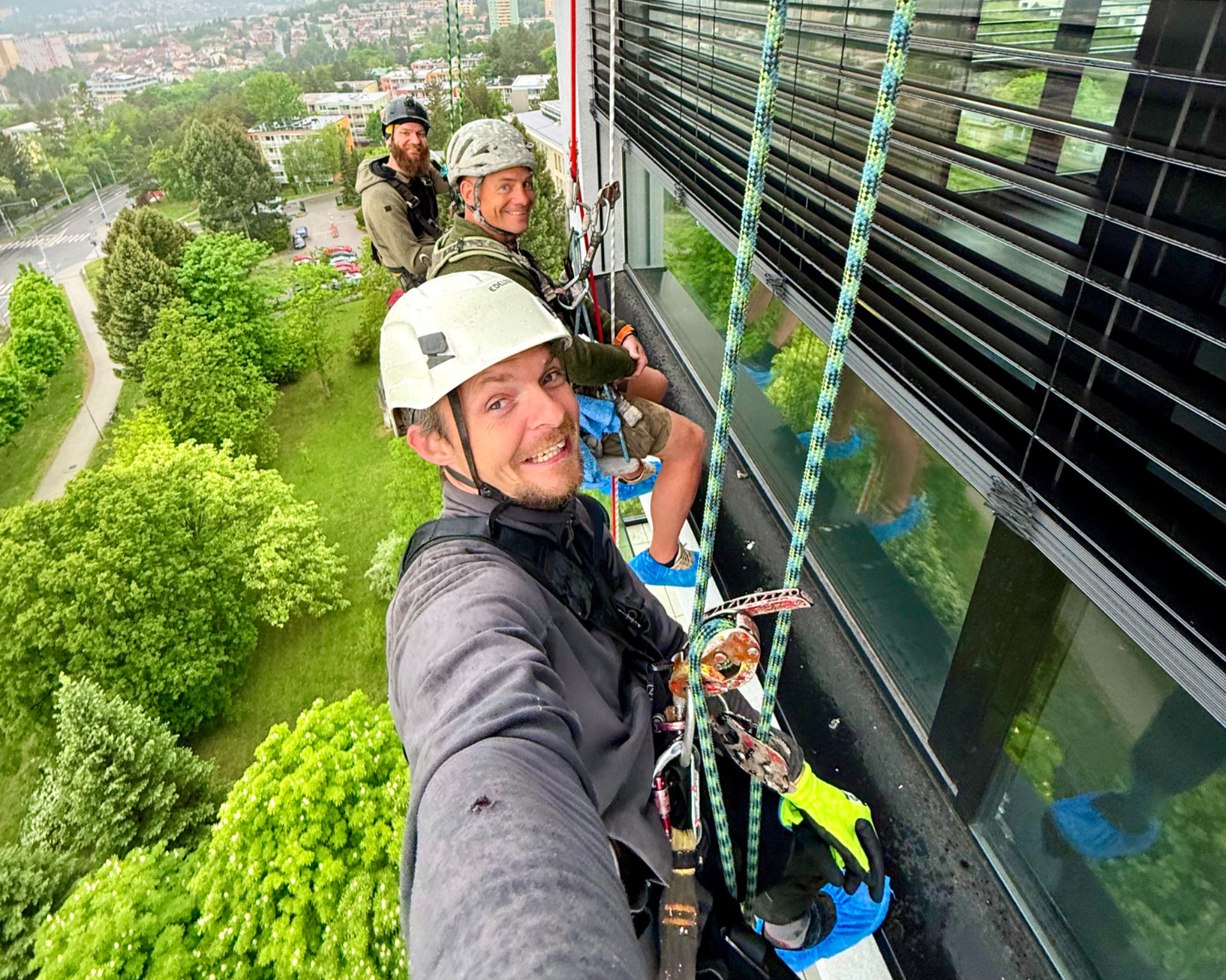 Two workers in helmets rappel beside a tall building, smiling over a green cityscape below.